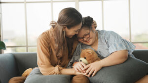 Mother supporting sweet down syndrome daughter to learn and relax from the internet, embracing child to encourage down syndrome girl at home.
